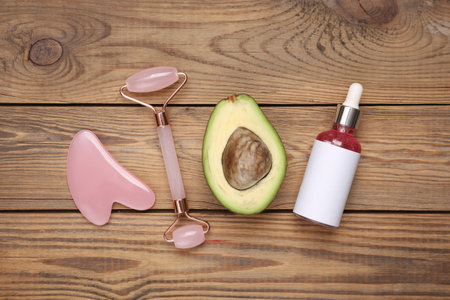 Face roller and gua sha stone massager, bottle of serum with avocado on wooden table. top viewの写真素材