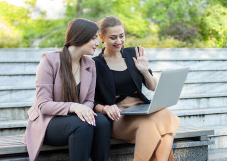 Two business women have a video call while sitting on a bench with laptop in the park