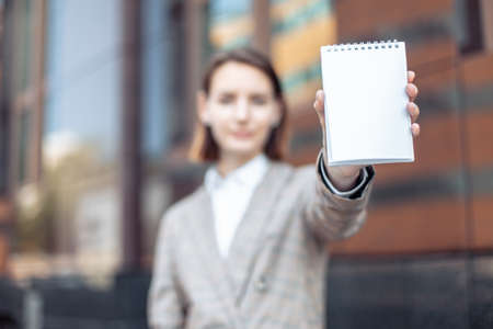 Young business woman shows a blank sheet of notepad on the background of a business building. focus on notepadの写真素材
