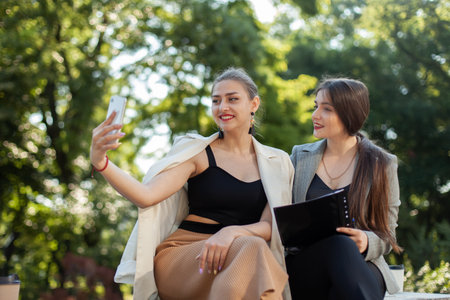 Two young business women make selfie on the phone while sitting on a bench in the parkの写真素材