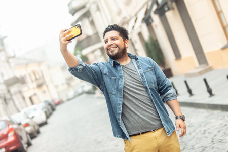 Young cheerful hispanic man in jeans jacket makes selfie on phone in european streetの写真素材