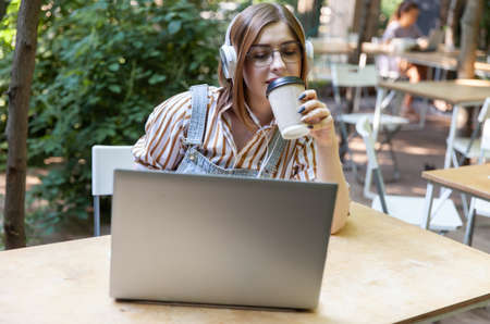 Young pretty woman with laptop and headphones drink coffee in outdoor coworking space