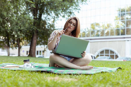 Pensive woman student looks into a laptop while sitting on a blanket in the park