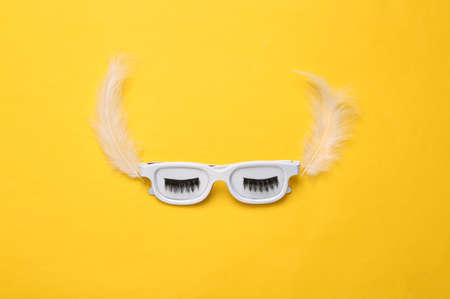Glasses with eyelashes and feathers on yellow background. Minimalism beauty concept. creative layout. top viewの写真素材