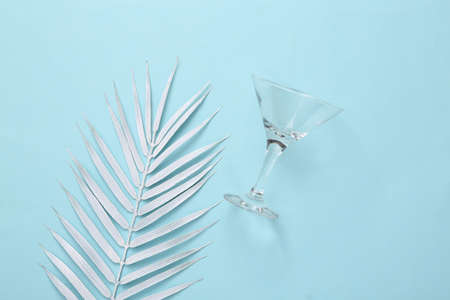 Cocktail empty glass with palm leaves on blue pastel background. Minimalism party concept. top view