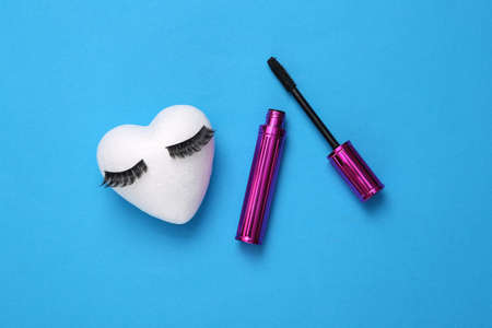 False eyelashes with heart and eyelash brush with mascara on blue background. Creative flat lay,...