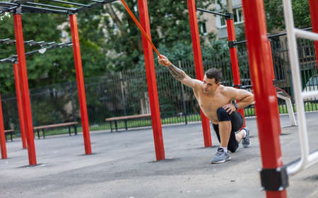 Athletic male athlete with a naked torso trains with a fitness elastic band on the workout site....