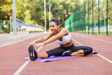 Young athletic woman practicing stretching while sitting on mat in the middle of stadium