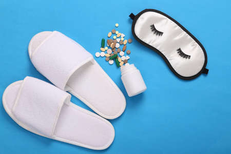 treatment of insomnia. Sleep mask with closed eyes, eyelashes, slippers and bottle of pills on blue...