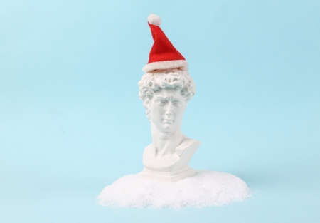 Plaster bust of David with Santa hat in snowdrift on turquoise background. Minimal Christmas layout