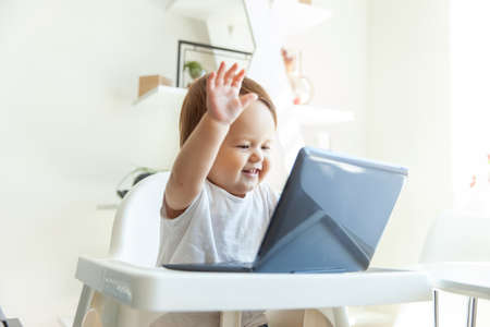 Little funny baby waving at the laptop screen while sitting at the children's table at homeの写真素材
