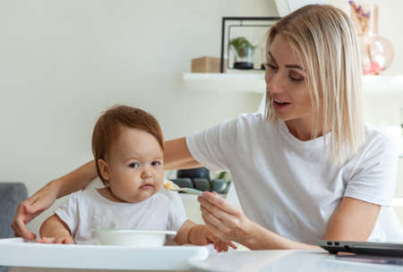A caring mother feeds her baby from a spoon while sitting at a table