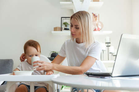 A young mother gives a drink to a small child from a cup while sitting at a table with a laptop. Maternal care conceptの写真素材
