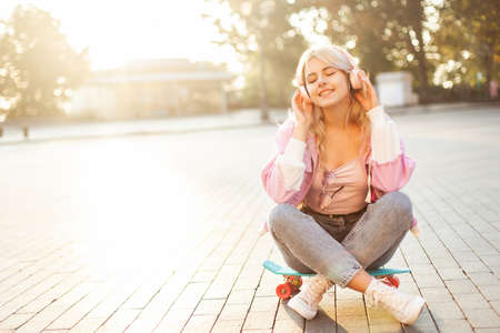 Young smiling woman enjoying music with headphones while sitting on a skateboard in the cityの写真素材
