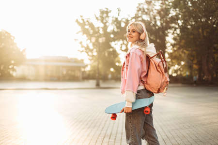 Young cool hipster girl in headphones with skateboard and backpack spends time in the cityの写真素材