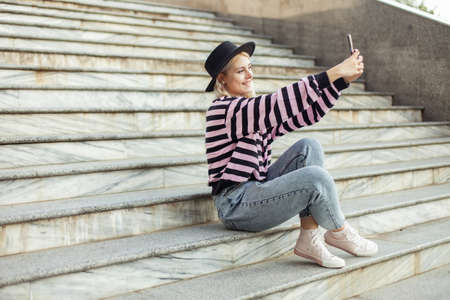 Young charming hipster girl in a hat makes a selfie on the phone while sitting on the stairs...