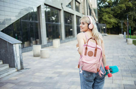 Young stylish girl in headphones with backpack and skateboard in the city