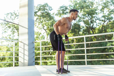 Athletic man trains muscles with fitness rubber bands outdoors. Muscular male with a naked torso...