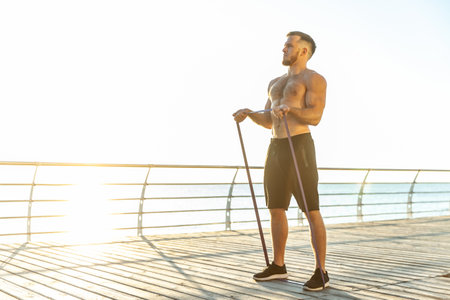 Young athletic man with a naked torso works out with fitness rubber bands in the early morning on...