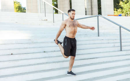 Young male athlete with a naked torso kneads the muscles doing stretching exercise while standing on...