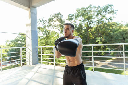 Muscular man training punches with boxing gloves outdoors