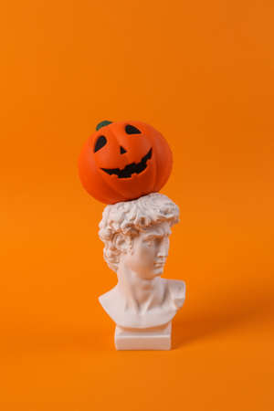 Plaster bust of David with jack lantern pumpkin head on orange background. Halloween concept. Minimal still life photoの写真素材