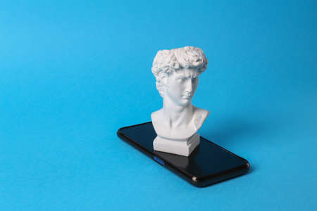 Antique bust of david with smartphone on blue background. Minimal still life.