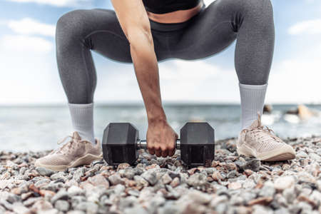 Strong athletic woman exercising with heavy dumbbell on the beach during the day with blue sky and...