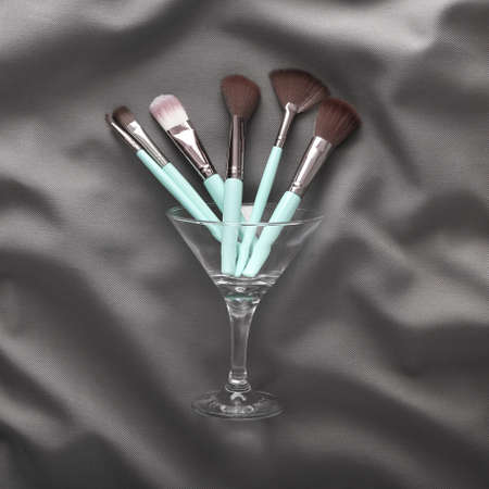 Cocktail glass with makeup brushes on gray crumpled silk background. party conceptの写真素材