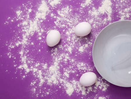 Culinary composition with flour, eggs and a plate on a purple background. top view