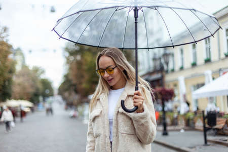 Cute fashion blonde woman in yellow sunglasses with a transparent umbrella walks along the city street. lifestyleの写真素材