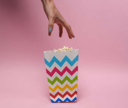 Woman's hungry hand reaching for popcorn box on pink background