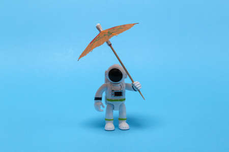 Astronaut in spacesuit figurine with umbrella on blue background.