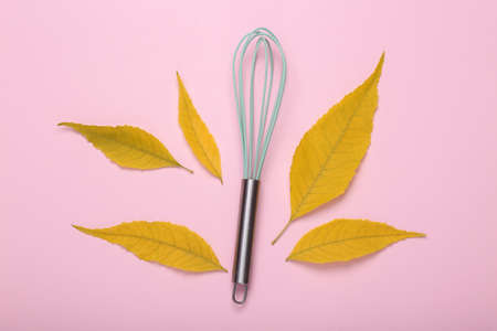 Kitchen whisk with fallen yellow leaves on a pink background. autumn time. top view