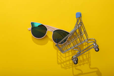 Shopping trolley with sunglasses on a yellow background with shadow. beauty and fashion