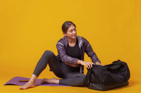 Smiling athletic woman in sportswear sits on mat and open sport bag on a yellow background. healthy lifestyleの写真素材