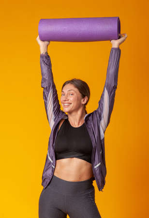 Cool cheerful athletic woman with yoga mat over her head on a yellow background. healthy lifestyle