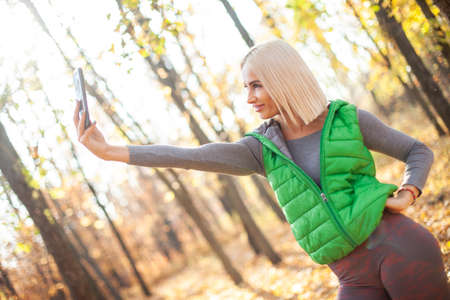 Smiling fitness blonde woman makes selfie on phone in autumn forest