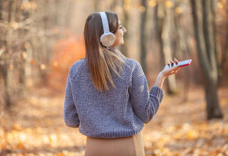 Portrait of a cute caucasian woman in a warm sweater listens to music in stereo headphones in the autumn forestの写真素材