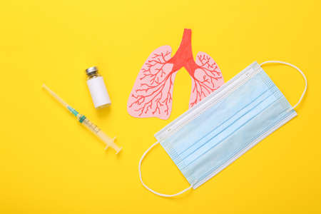 Vaccine bottle with syringe, medical mask and anatomical lungs on a yellow background. Pandemic...