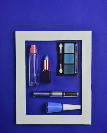 Female cosmetics in a white frame on blue background. Shadows for make-up, lipstick, nail polish, perfume bottle. minimalist trend. top view. copyspace.の写真素材