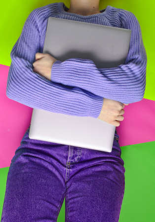 A girl in jeans and a sweater lies on a colored paper background and hugs a laptop. top view.