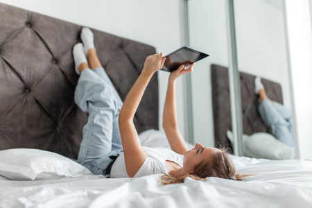 Young beautiful caucasian woman with a tablet lying on a bed in bright roomの写真素材