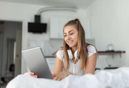 Young smiling woman with headphones looks into the screen of the tablet while lying on the bed with a bright roomの写真素材