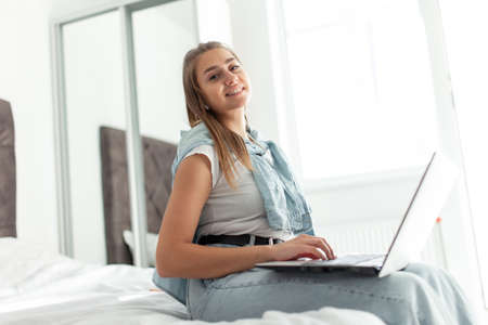 Young smiling woman using laptop while sitting on bed in bright room