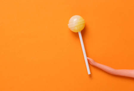 Doll hand holding lollipop on orange background. Minimal halloween, junk food, sweets, concept. Top...