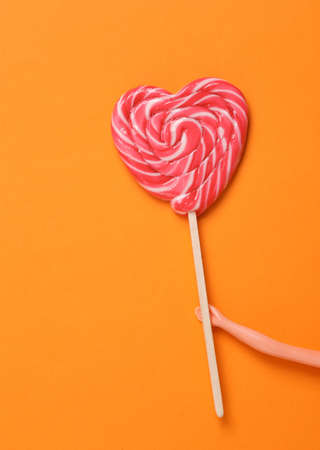 The doll's hand holds a heart-shaped lollipop on an orange background. Minimal still lifeの写真素材