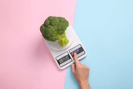 Hand presses the button of kitchen scale with broccoli on a blue-pink pastel background. Weight loss conceptの写真素材