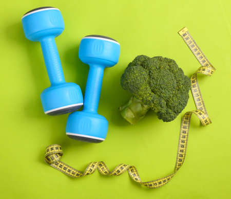 Broccoli with a measuring tape and dumbbells on a green background. Weight loss and fitness concept. Healthy food. Top viewの写真素材