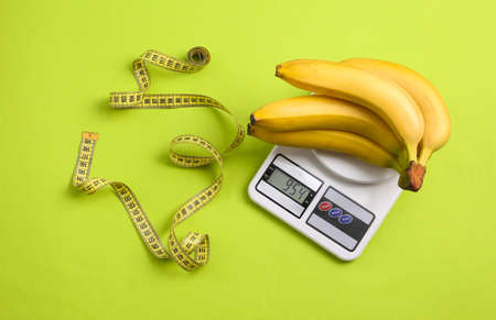 Kitchen scales with bananas and measure tape on a green background. Slimming, weight loss concept, healthy foodの写真素材